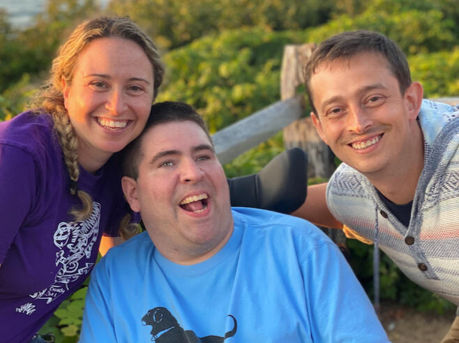 Three people smile at the camera, a woman with blonde hair in a braid on the left, a man in a wheelchair in the middle, and a man on the right with light brown hair.